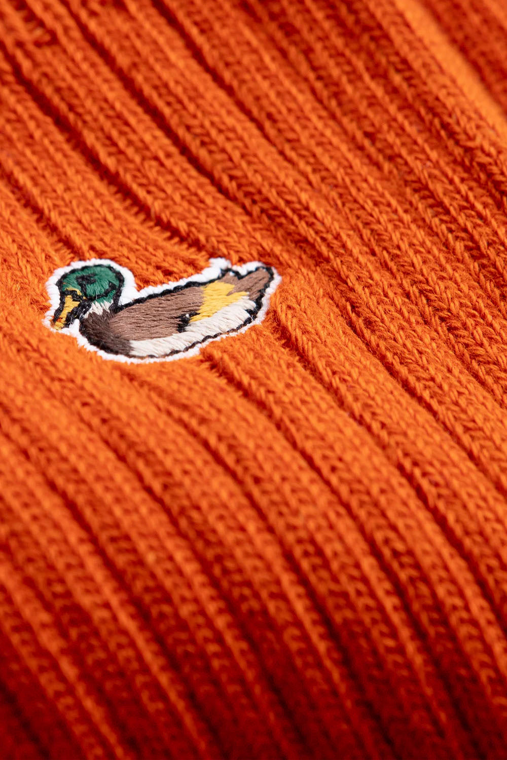 Close-up of a ribbed orange knit fabric with a small embroidered mallard duck patch on the left side.