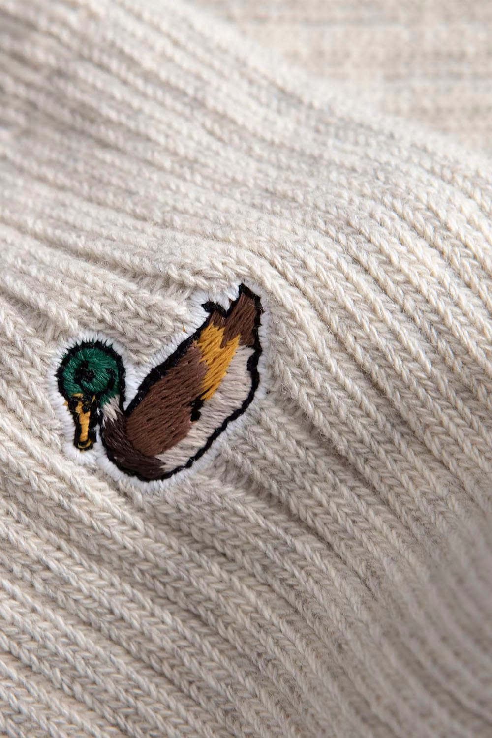 Close-up of a ribbed cream-colored knit fabric, likely a sock, featuring an embroidered mallard duck motif with a green head, brown body, and yellow accents.