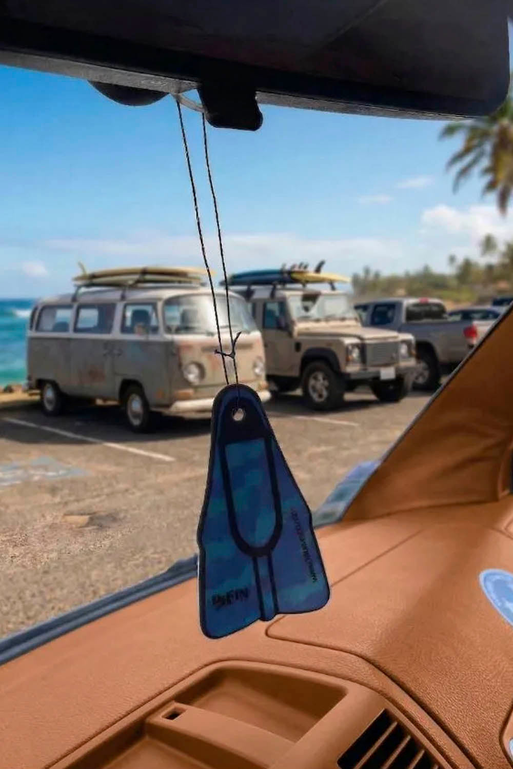 Car air freshener hanging from a car's rearview mirror with a beach and vehicles in the background.