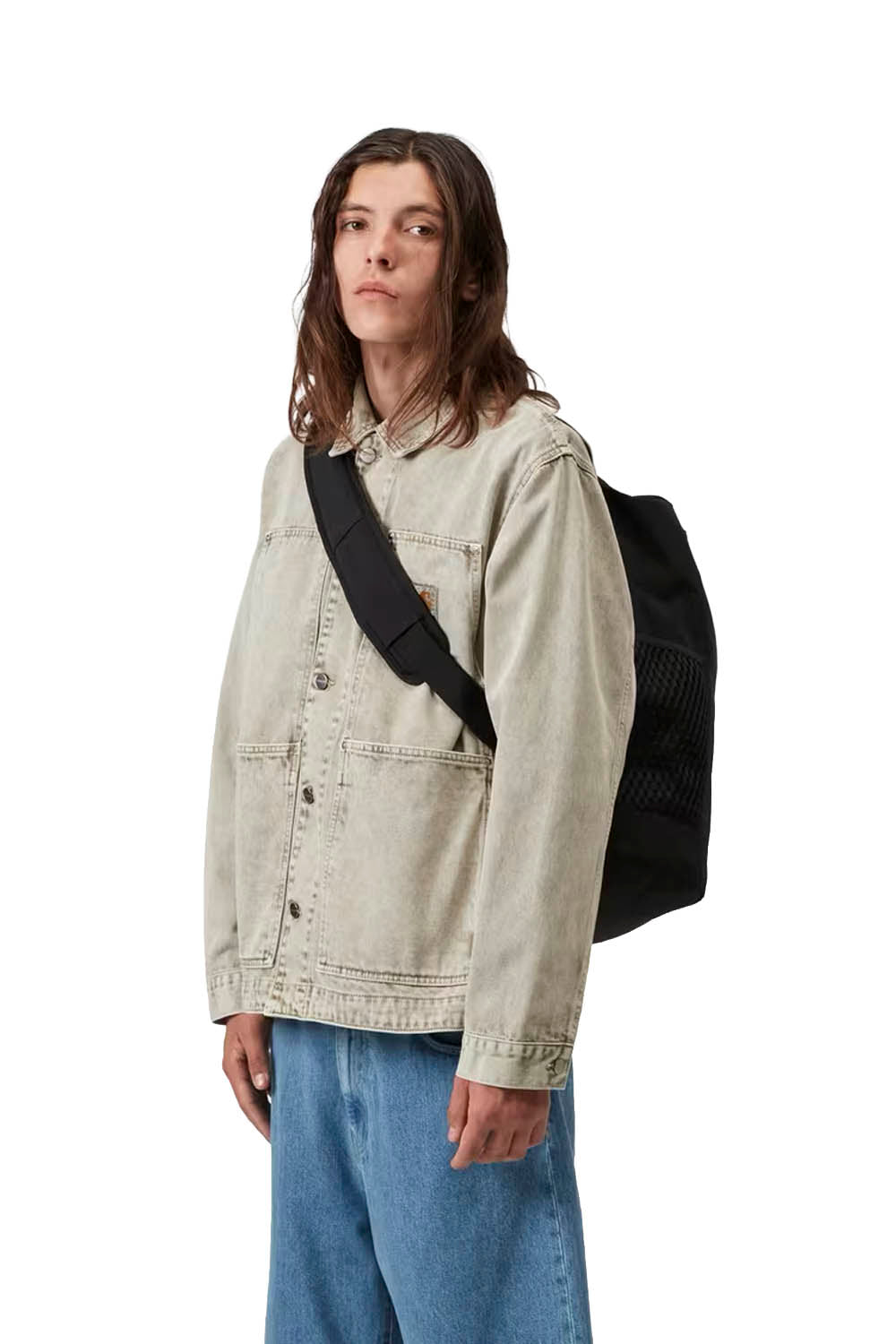Person wearing a beige jacket and blue jeans with a black crossbody backpack on a white background