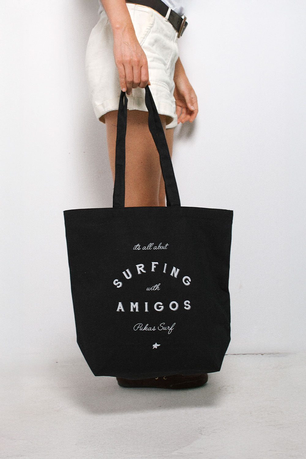 A person wearing white shorts holds a large black canvas tote bag with white printed text that reads 'SURFING with AMIGOS' and 'Pukas Surf'.