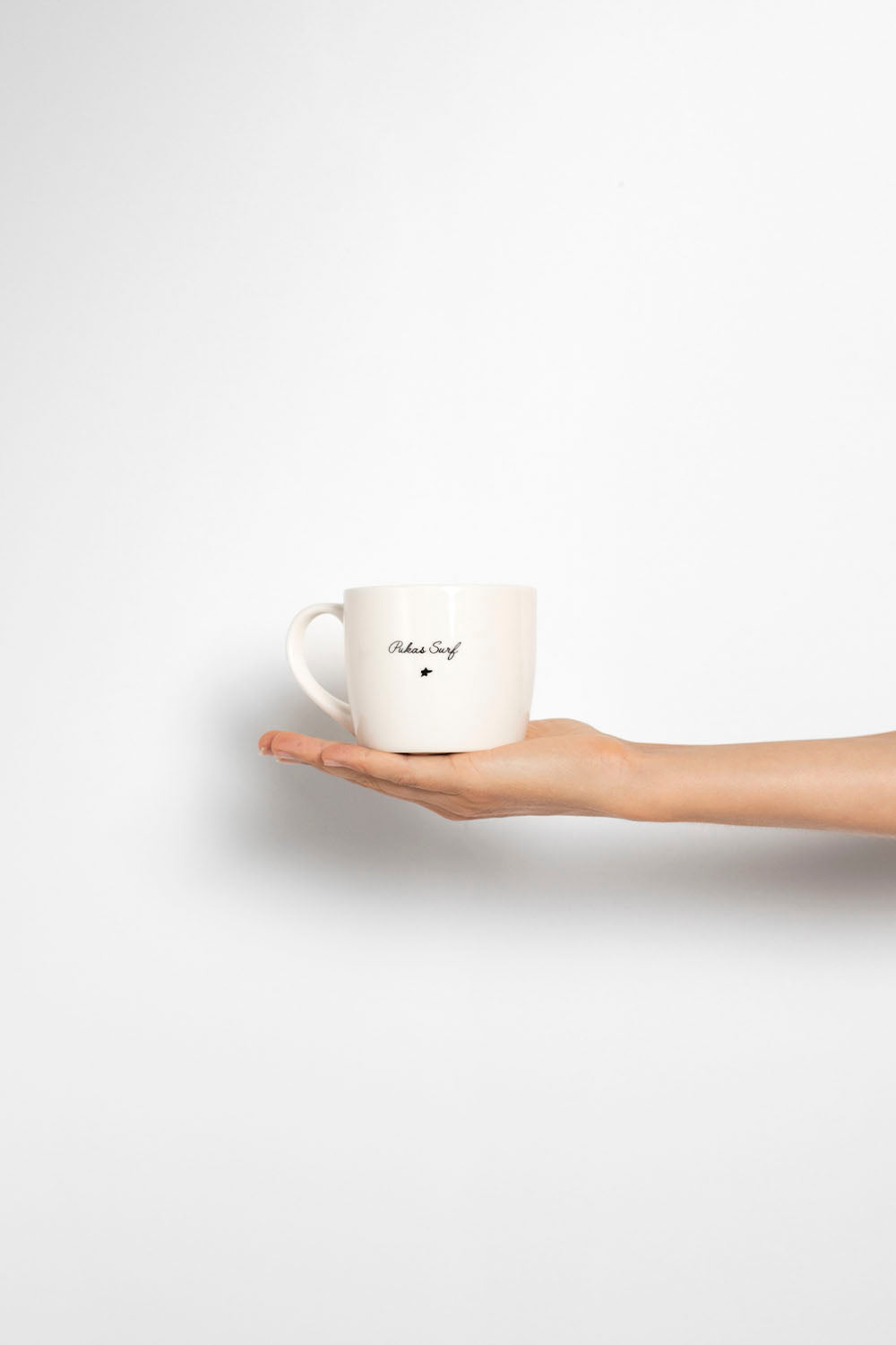 A hand holding a white ceramic mug with black text that reads 'PUKAS SURF'  against a clean white background.