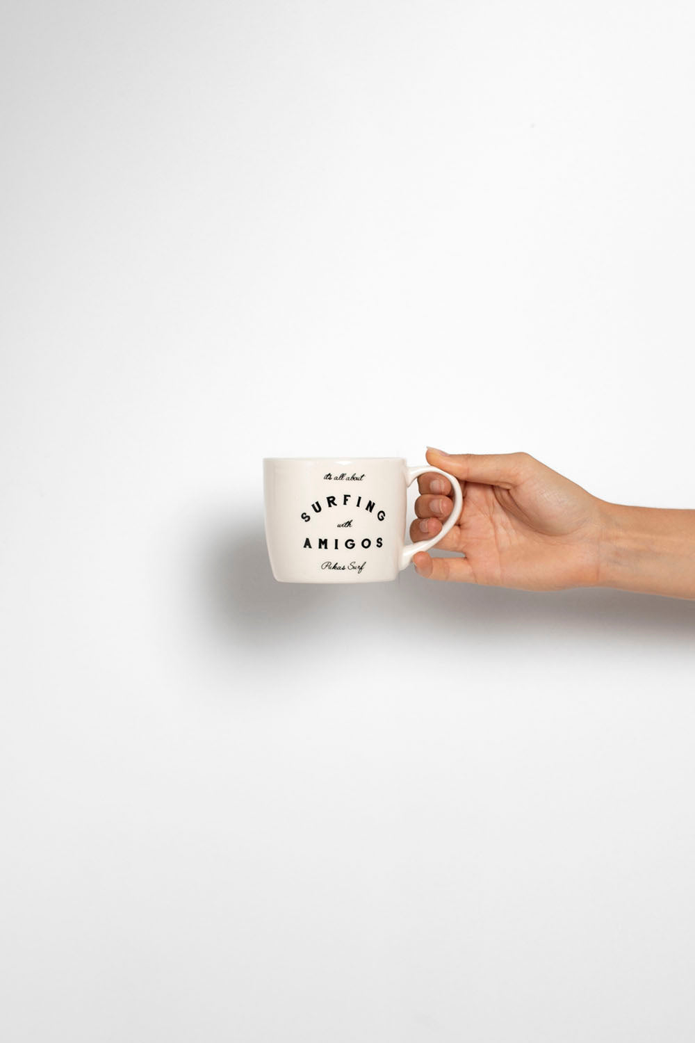 A hand holding a white ceramic mug with black text that reads 'SURFING AMIGOS'  against a clean white background.