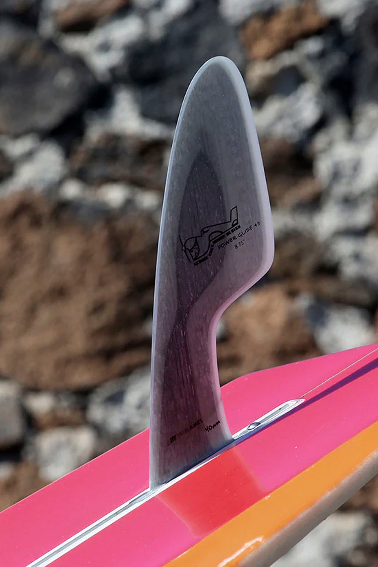 Close-up of a pink and orange surfboard with a sigle fin on a rocky background
