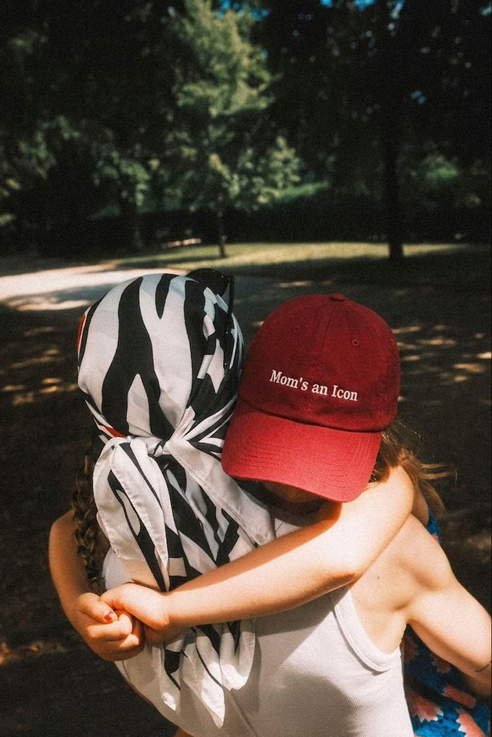 A person outdoors wearing a red baseball cap with the embroidered text 'Mama's an Icon' and a black-and-white patterned headscarf, embracing another person in a park setting.