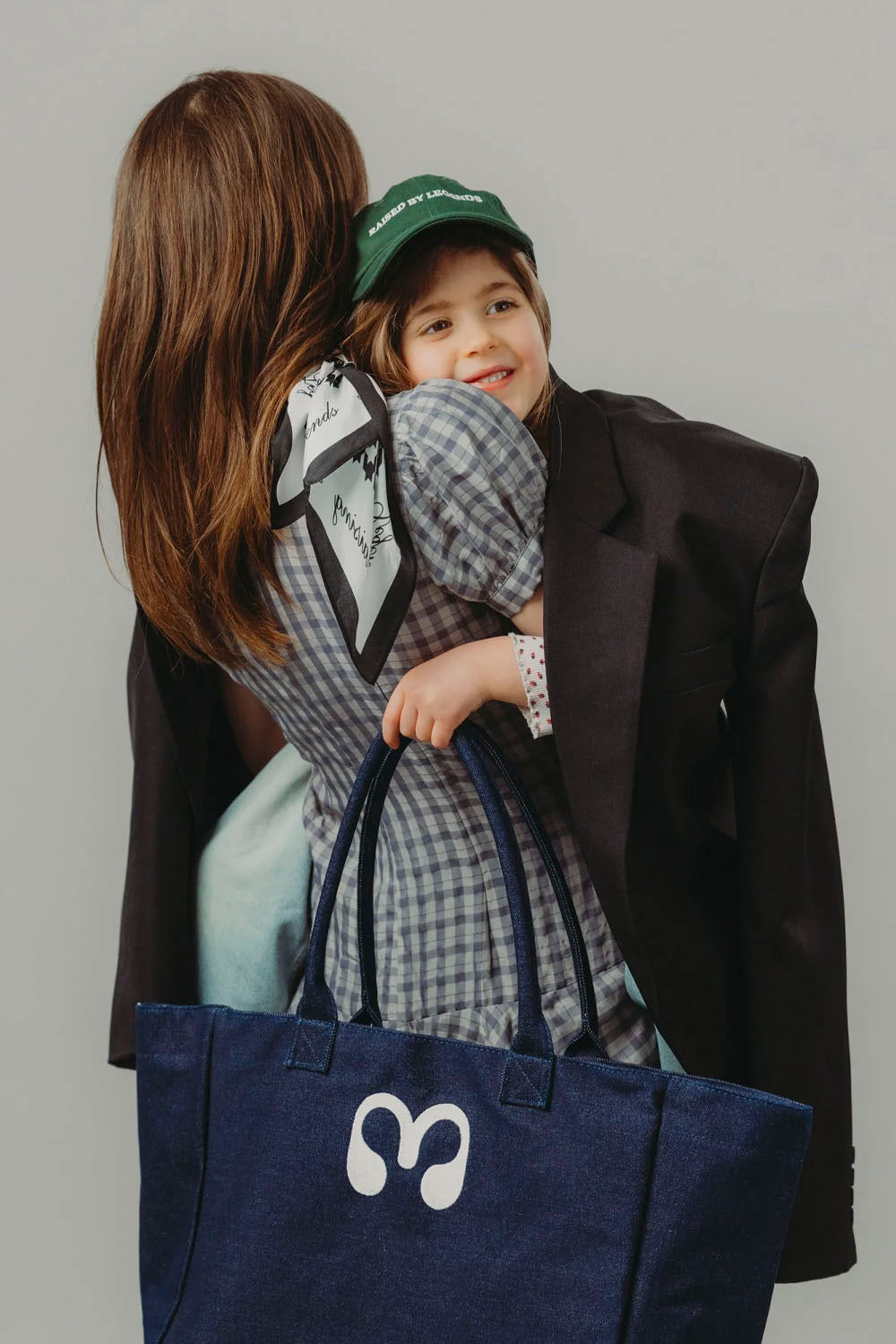 A woman in a black blazer and a young girl in a plaid shirt and green cap hug while holding a large navy blue tote bag with a white headphone logo. The background is a solid light gray studio wall.