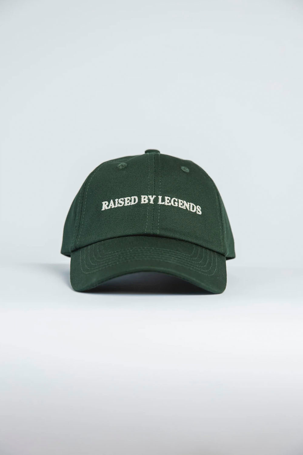 A dark green baseball cap with 'RAISED BY LEGENDS' embroidered in white across the front, shown against a light gray studio background.