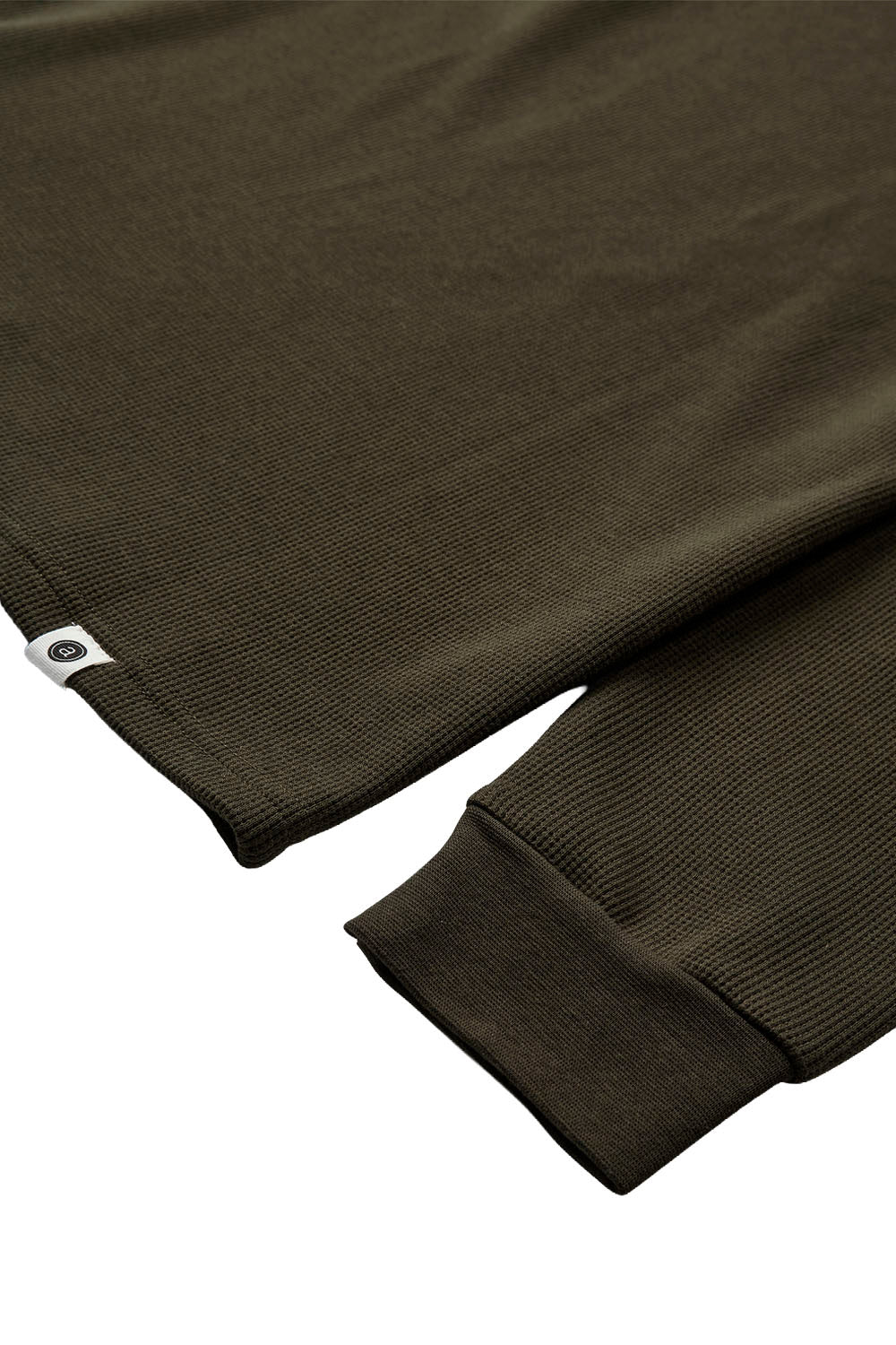 Close-up of a ribbed knit long-sleeve top in dark olive green, showing the sleeve cuff and a small white brand tag near the hem.