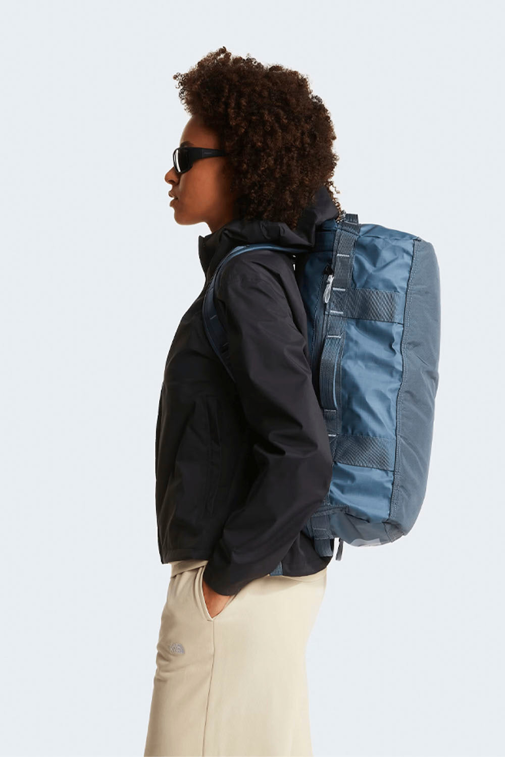 Person wearing a black jacket and beige pants with a blue backpack on a light gray background