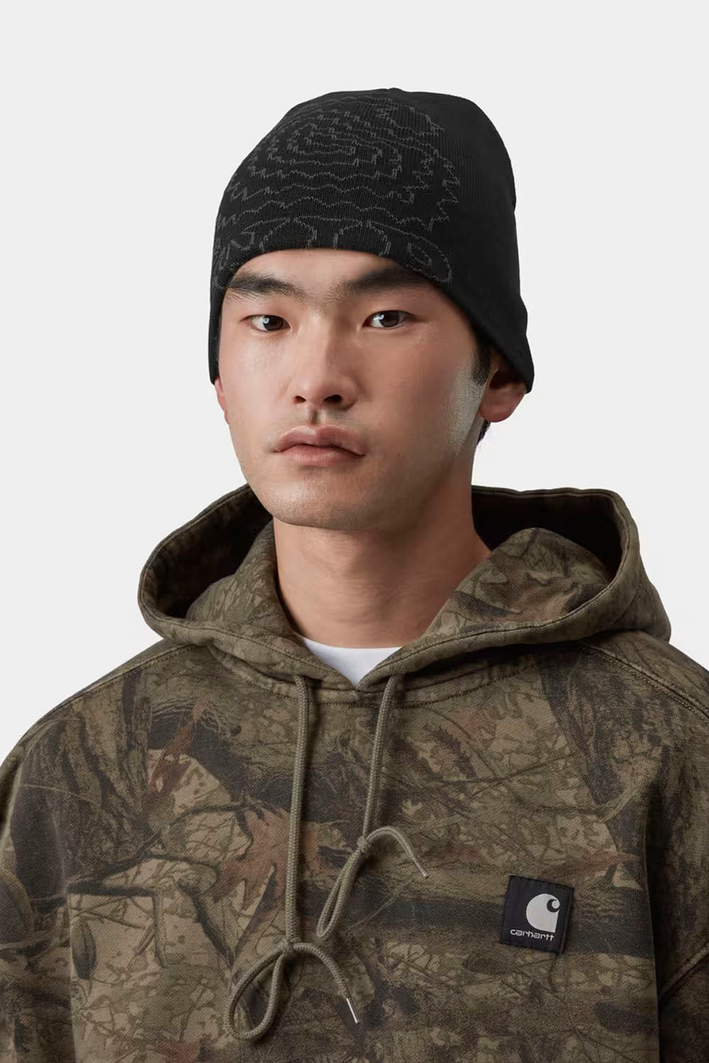 A man wearing a black beanie with a subtle line pattern and a camouflage hoodie with a visible Carhartt logo patch on the chest, set against a plain light gray background.