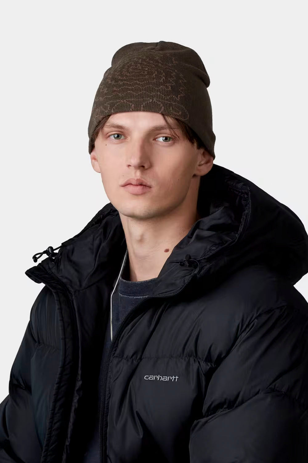A man wearing a dark brown knit beanie with a subtle pattern and a black Carhartt puffer jacket, photographed against a light gray studio background.