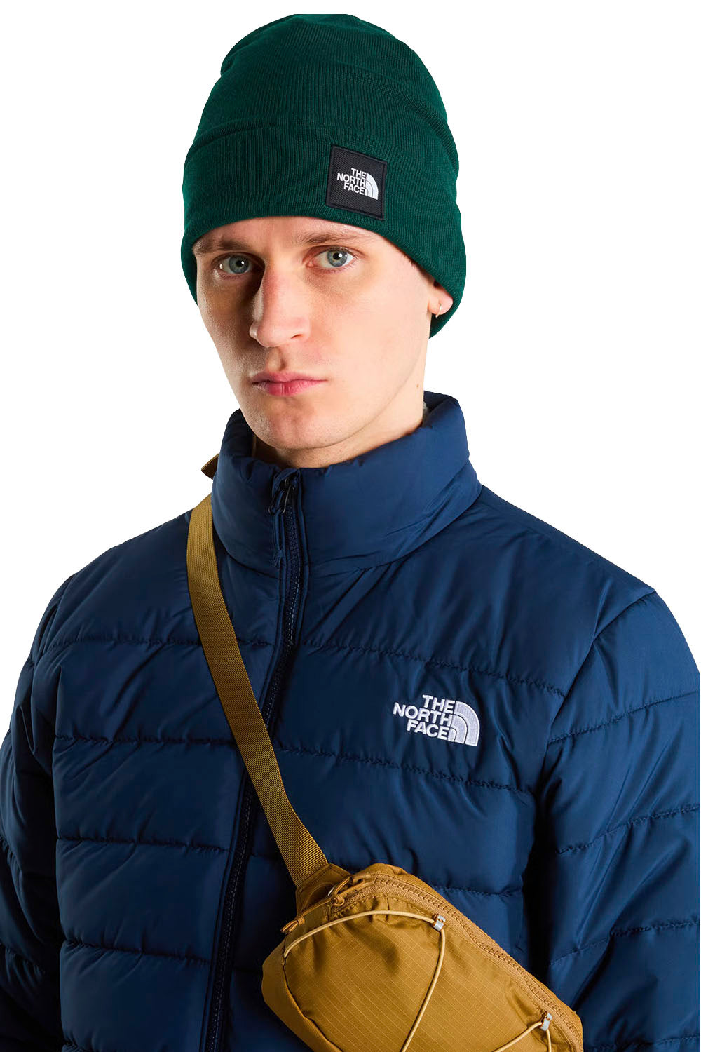 A model wearing a dark green beanie, a navy blue puffer jacket with The North Face logo, and a tan crossbody bag, photographed against a solid white background.