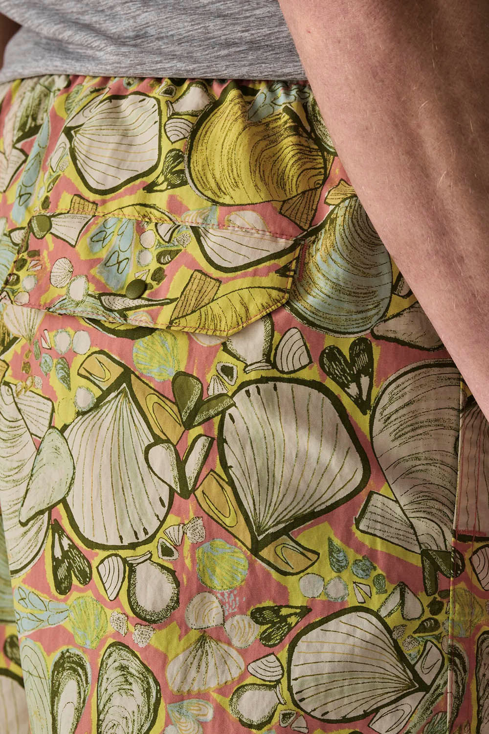 Close-up of a person wearing patterned shorts with a pink background and large shell illustrations in yellow, green, and gray, paired with a gray top.
