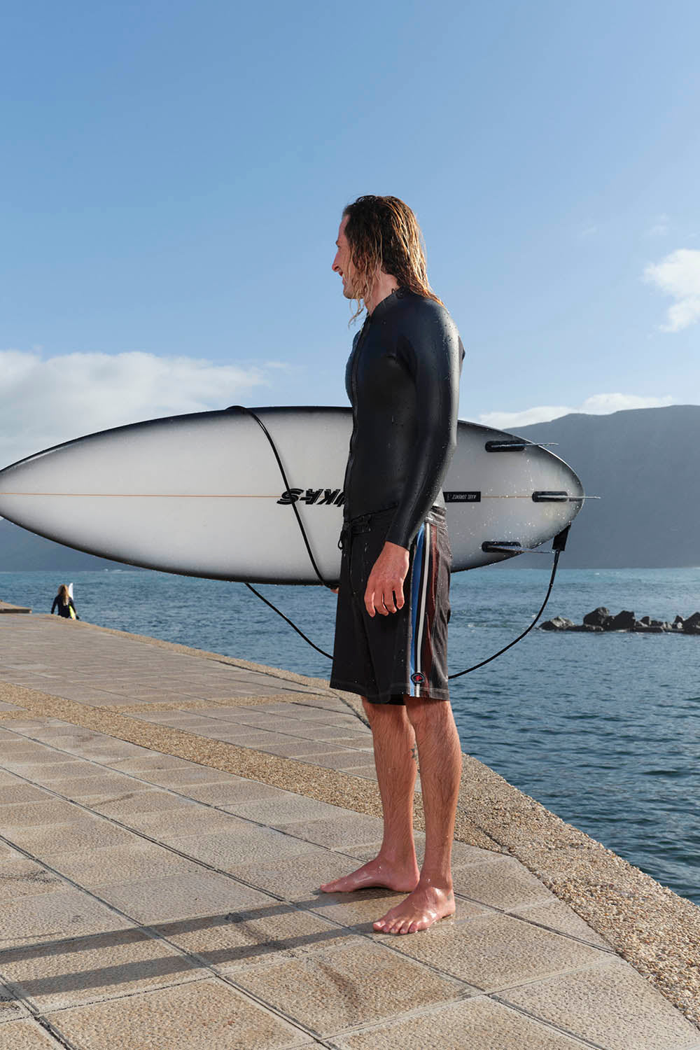 Man holding a surfboard by the water with mountains in the background