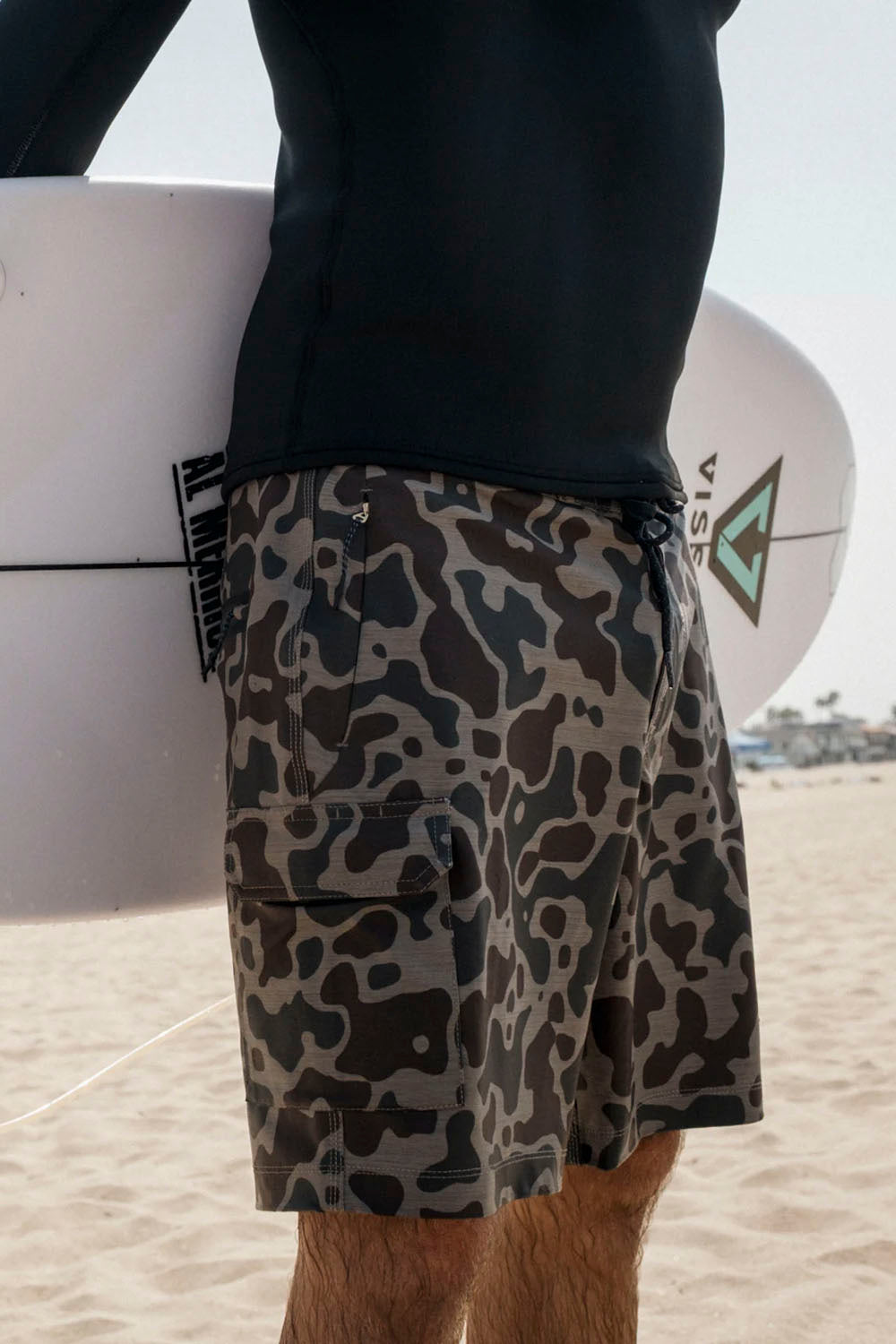 Person wearing camouflage shorts holding a surfboard on a beach