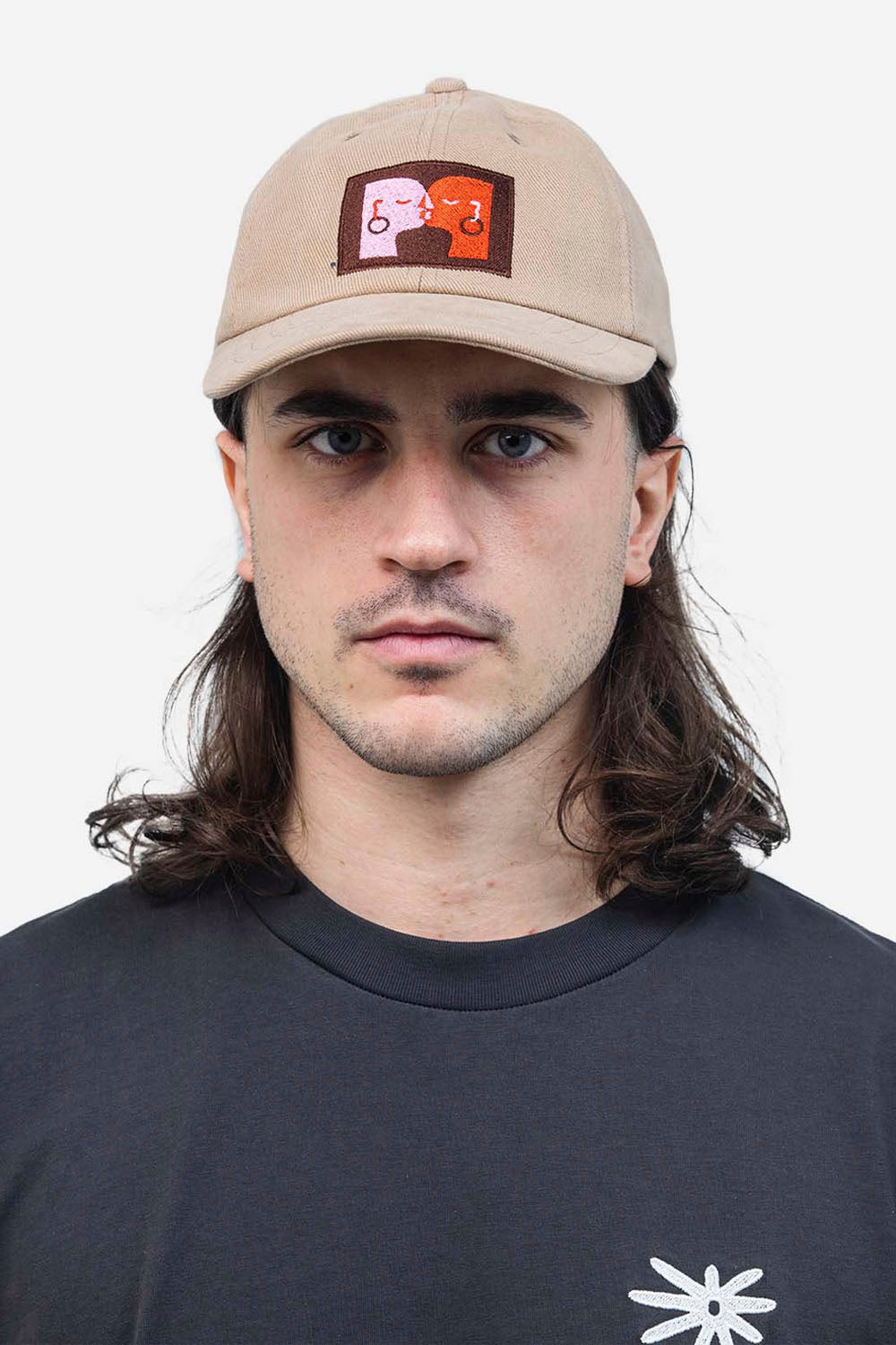 Person wearing a beige cap with a logo and a black t-shirt with a white logo on a plain background