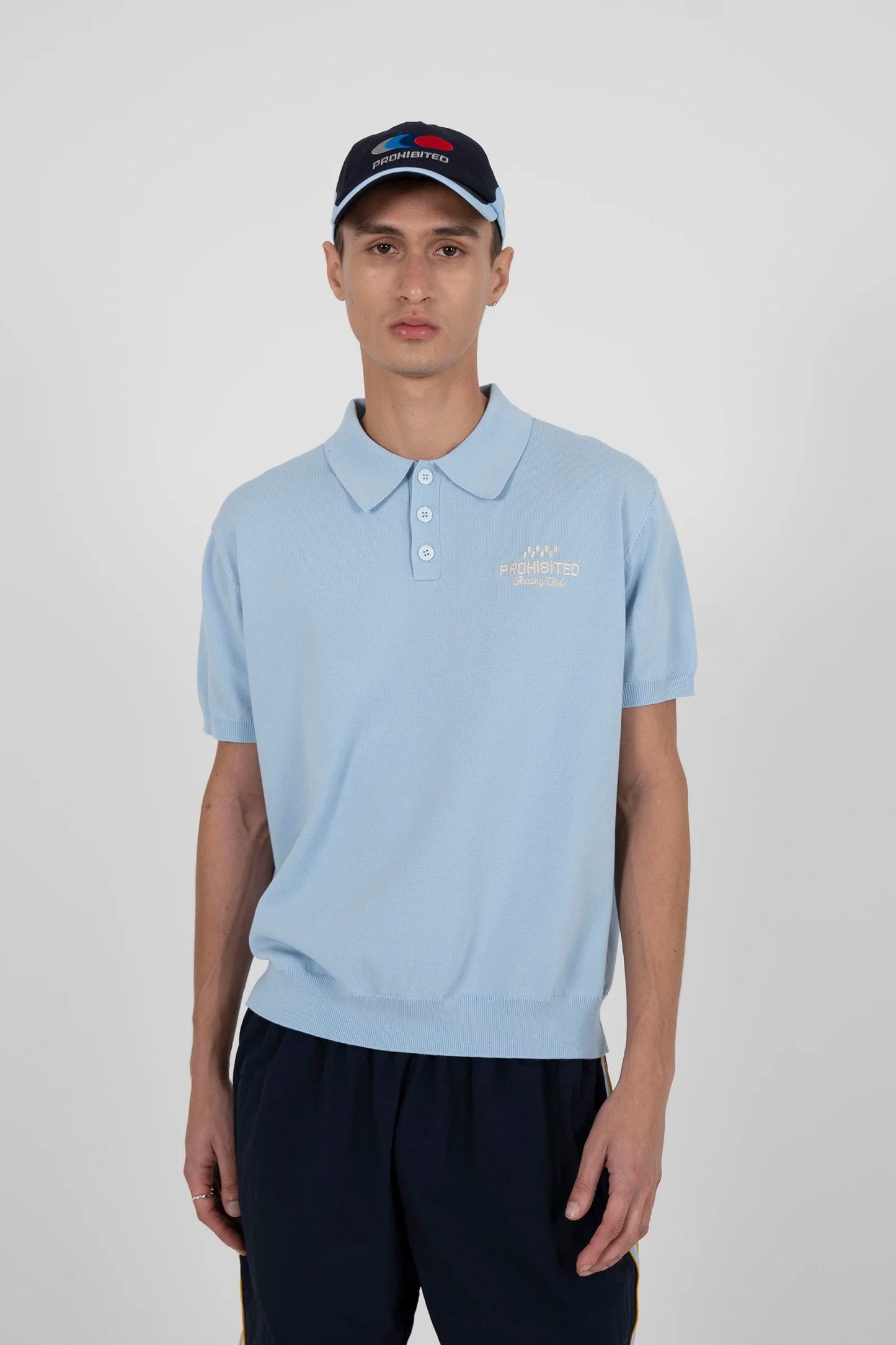 Man wearing a light blue polo shirt with a logo, dark pants and PROHIBITED tire racer cap on a white background