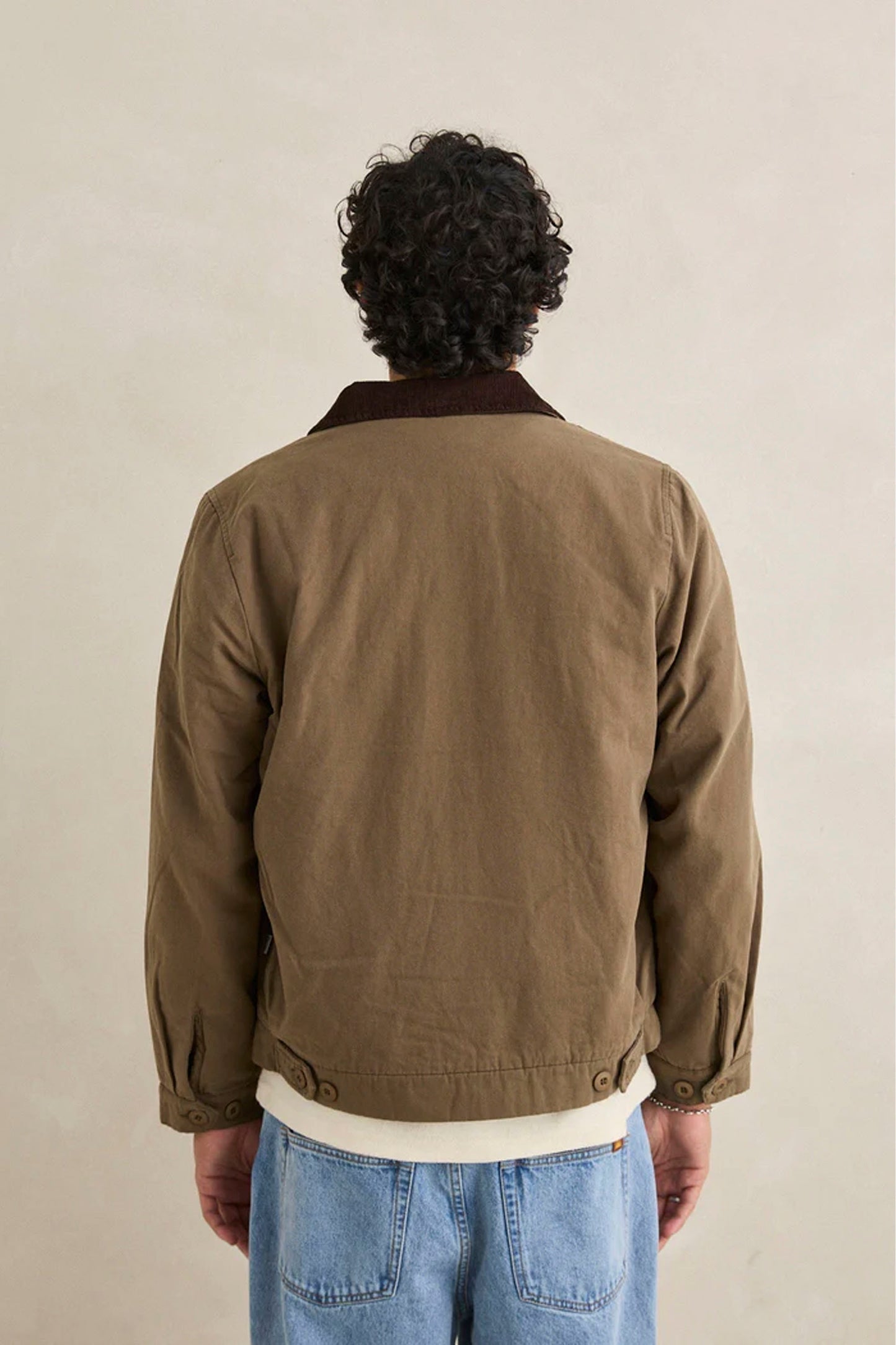 Man wearing Rhythm James Jacket oak, white shirt, blue jeans, and black shoes against a beige background