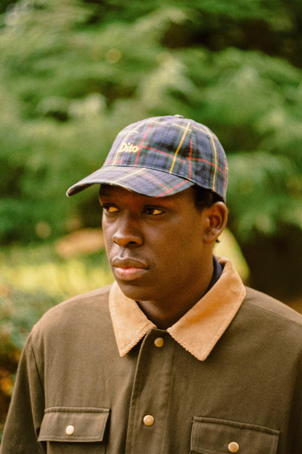 A man wearing a blue and yellow plaid baseball cap with 'boston' embroidered on the front, paired with a brown jacket featuring a tan corduroy collar, set against a blurred green outdoor background.