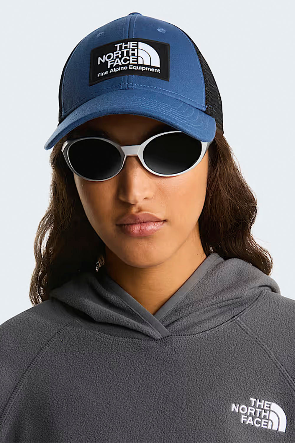 Person wearing a blue The North Face cap and sunglasses with a gray hoodie on a light gray background