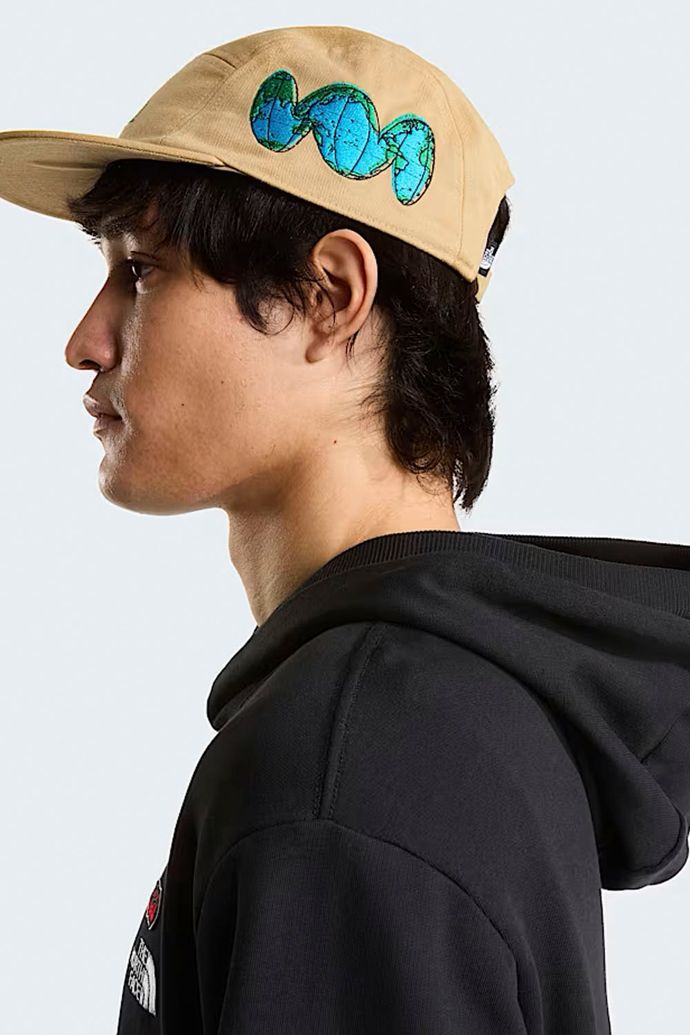 Person wearing a beige cap with a blue design and black hoodie on a light gray background