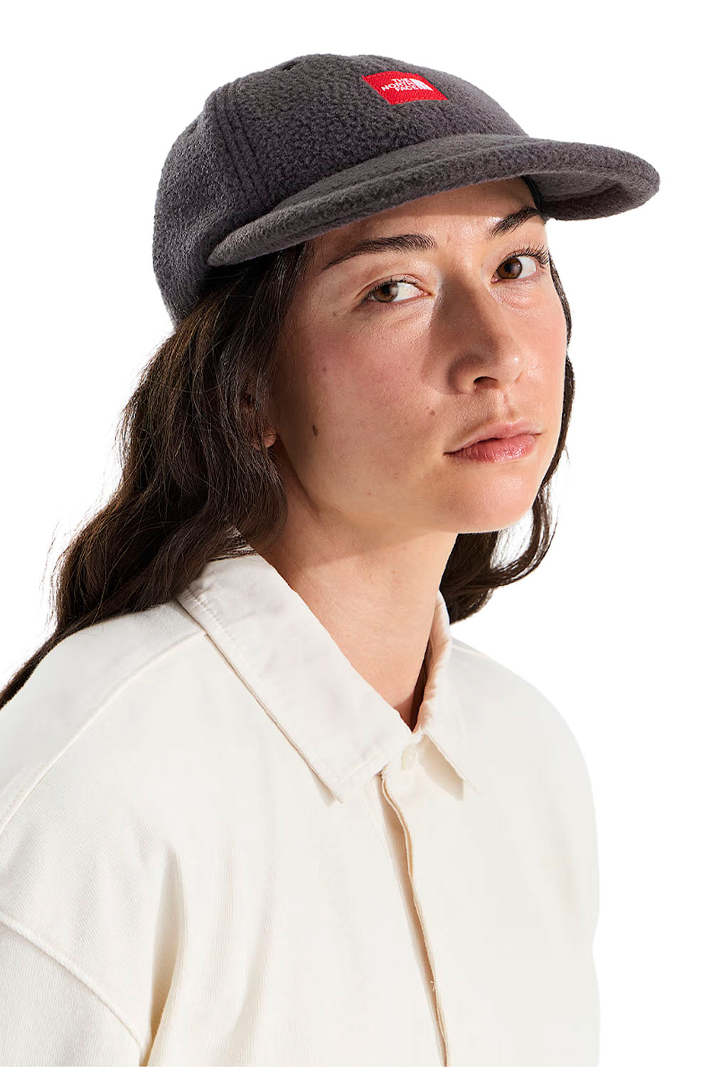 A woman wearing a dark gray fleece baseball cap with a small red rectangular logo patch on the front, paired with a cream-colored collared shirt, photographed against a solid white background.