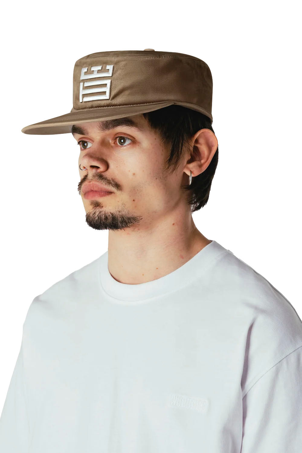 A man wearing a brown flat-brim cap with a white geometric logo on the front, paired with a white crewneck t-shirt, photographed against a solid white background.