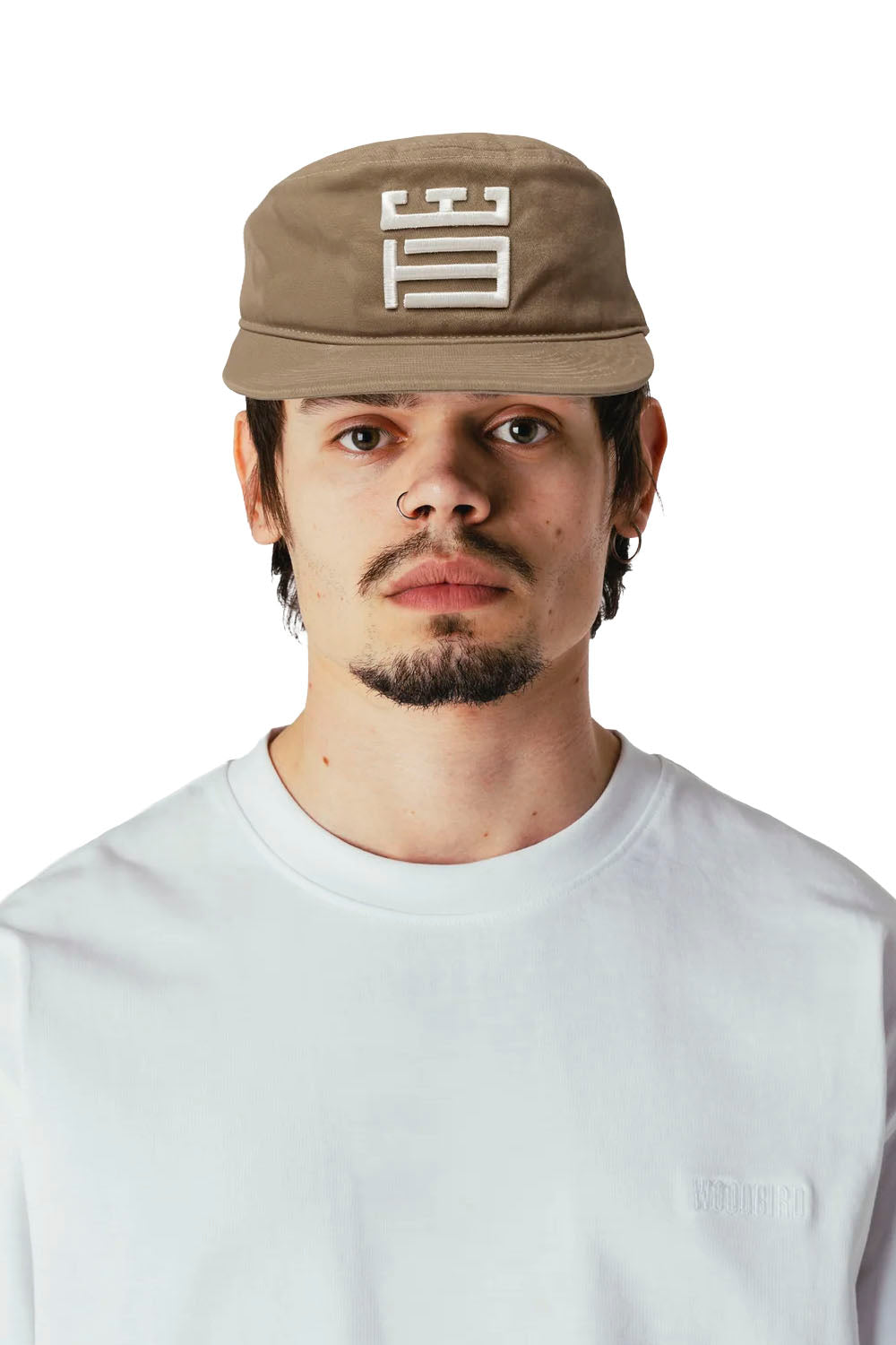 A man wearing a beige baseball cap with a white geometric logo and a white t-shirt, photographed against a solid white background.