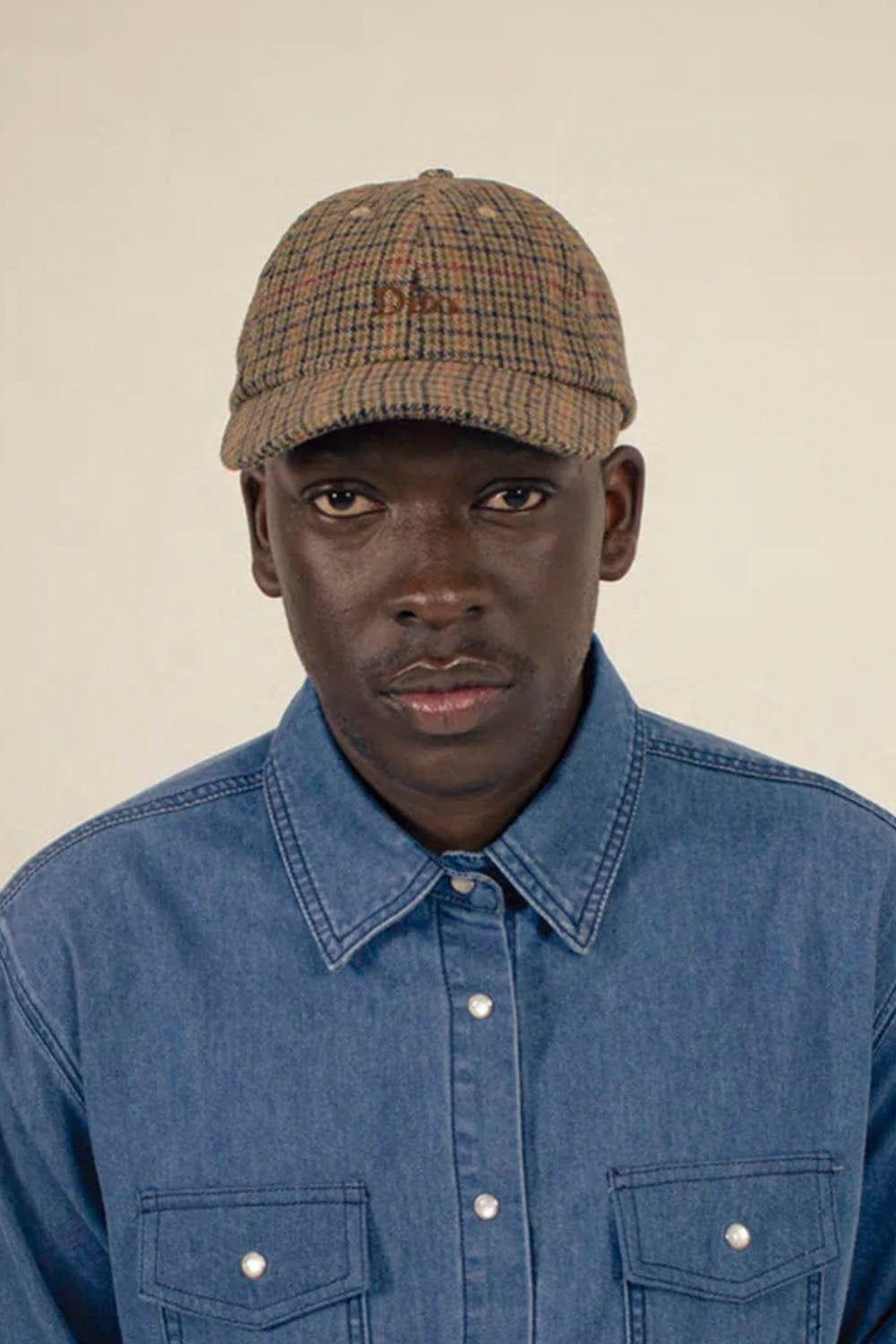 A man wearing a brown and beige plaid flat-brim cap with a small embroidered logo, paired with a blue denim button-up shirt, photographed against a solid light beige background.