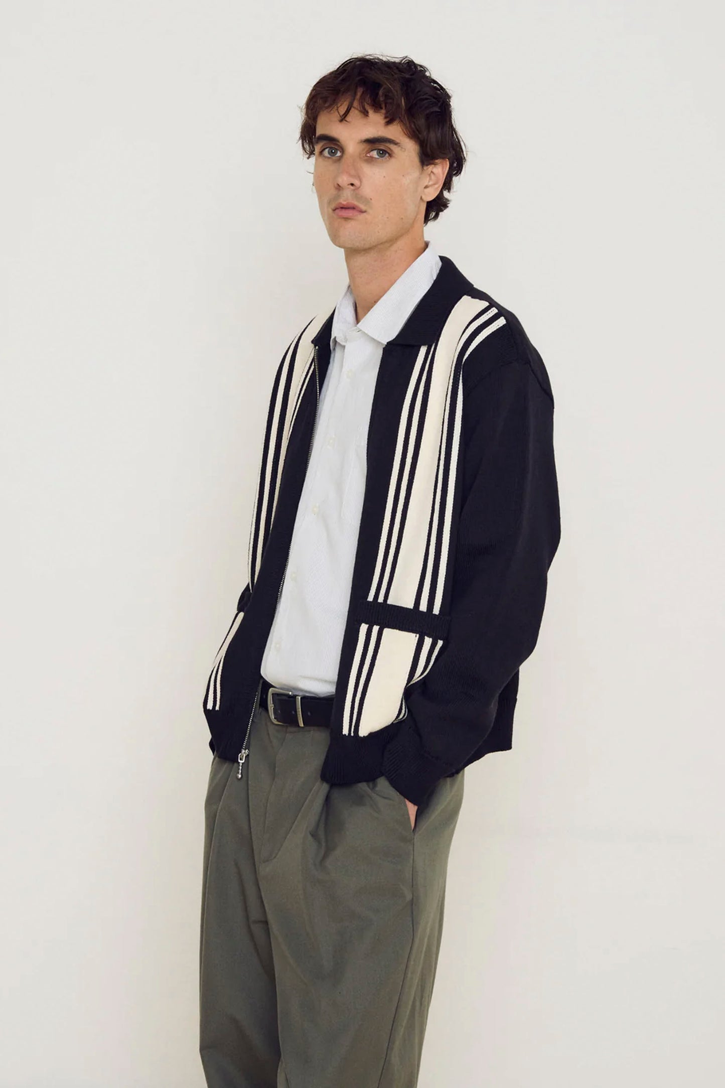 Man wearing a Black OBEY Matt Zip Up cardigan with white stripes over a light shirt and gray pants on a plain background