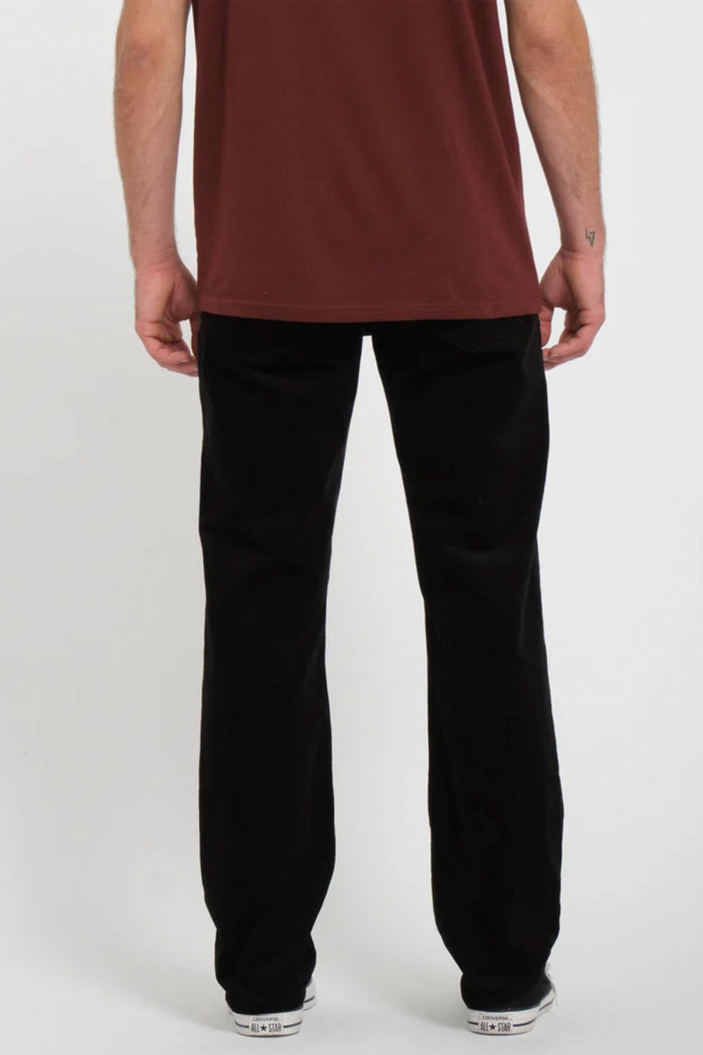 A person shown from the back wearing black straight-leg pants and a rust-brown t-shirt, standing against a light gray studio background.