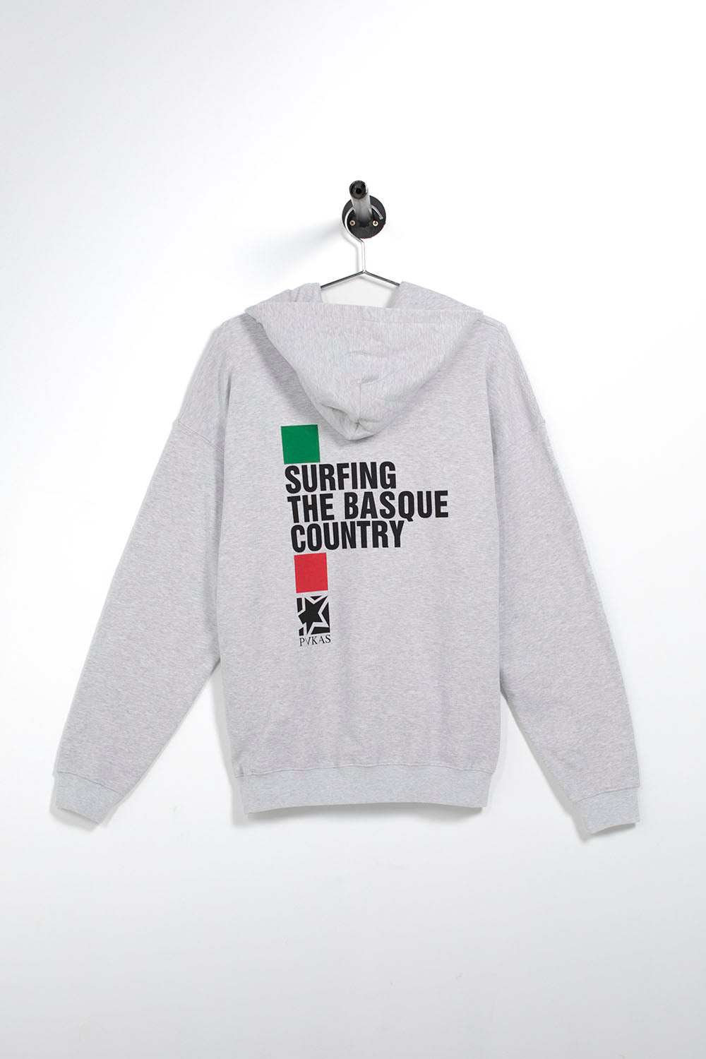 A light gray heather hoodie hanging on a black hanger against a white wall. The back of the hoodie features a graphic print with a green and red vertical flag design and the text 'SURFING THE BASQUE COUNTRY' alongside a small Basque emblem.