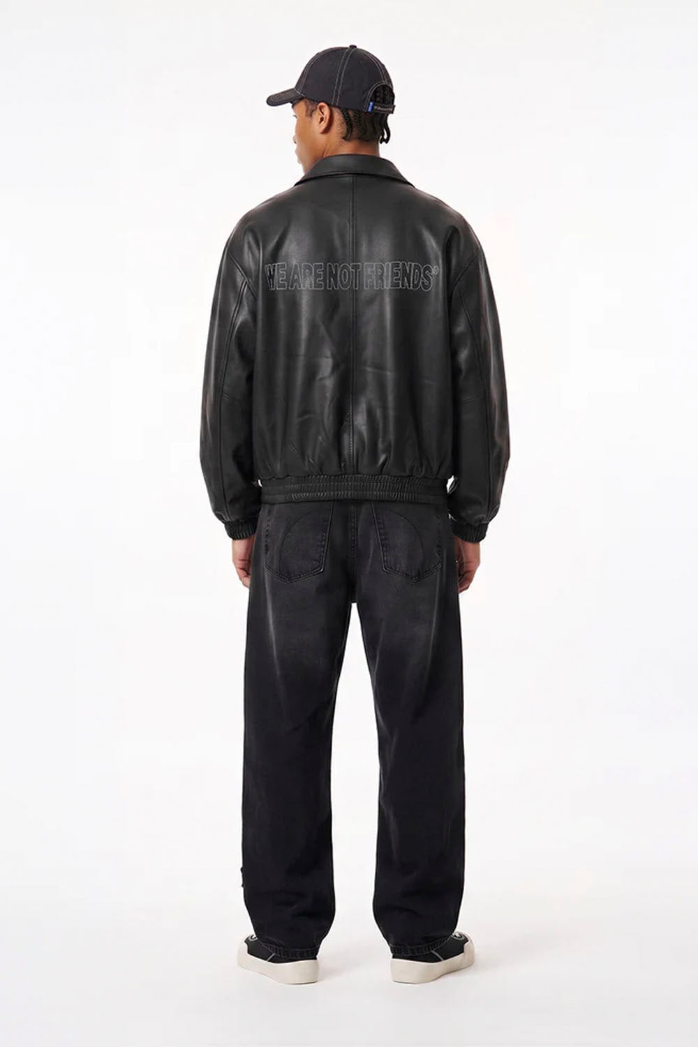 A man viewed from behind wearing a black leather jacket with 'THE TRAPERS' text on the back, black jeans, a black cap, and black sneakers, shot against a white studio background.