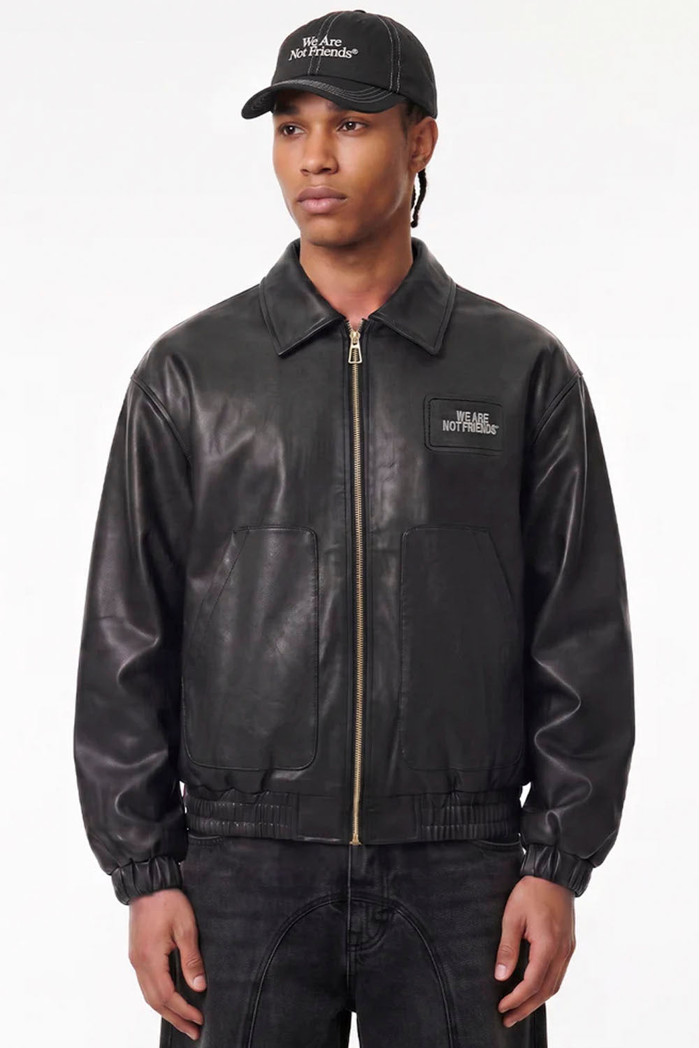 A model wearing a black leather bomber jacket with a gold zipper and a black baseball cap, both featuring the white embroidered text 'WE ARE NOT ANIMALS'.