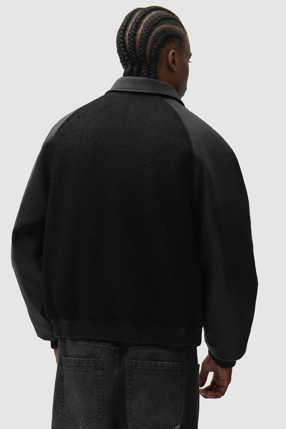 A man viewed from behind wearing a black knit sweater jacket with contrasting dark gray leather or faux-leather sleeves and collar, paired with dark jeans against a light gray studio background.