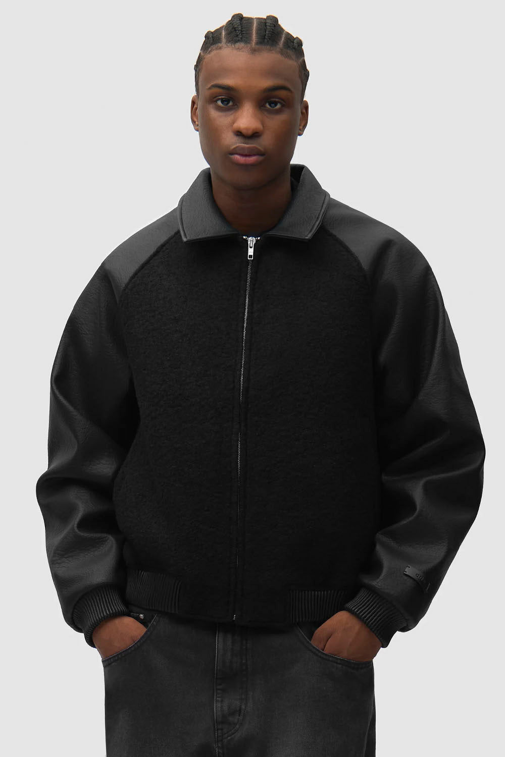 A man wearing a black zip-up jacket with contrasting black leather sleeves and collar, paired with dark gray jeans, against a solid light gray background.