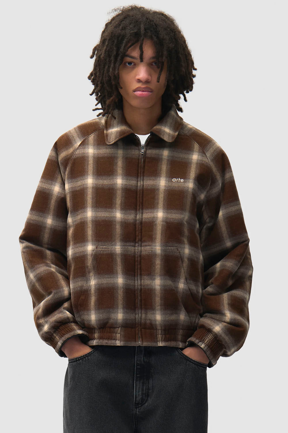 A model with dark curly hair wearing a brown and beige plaid fleece jacket with a small white 'art' logo on the chest, paired with dark gray jeans, against a solid light gray background.