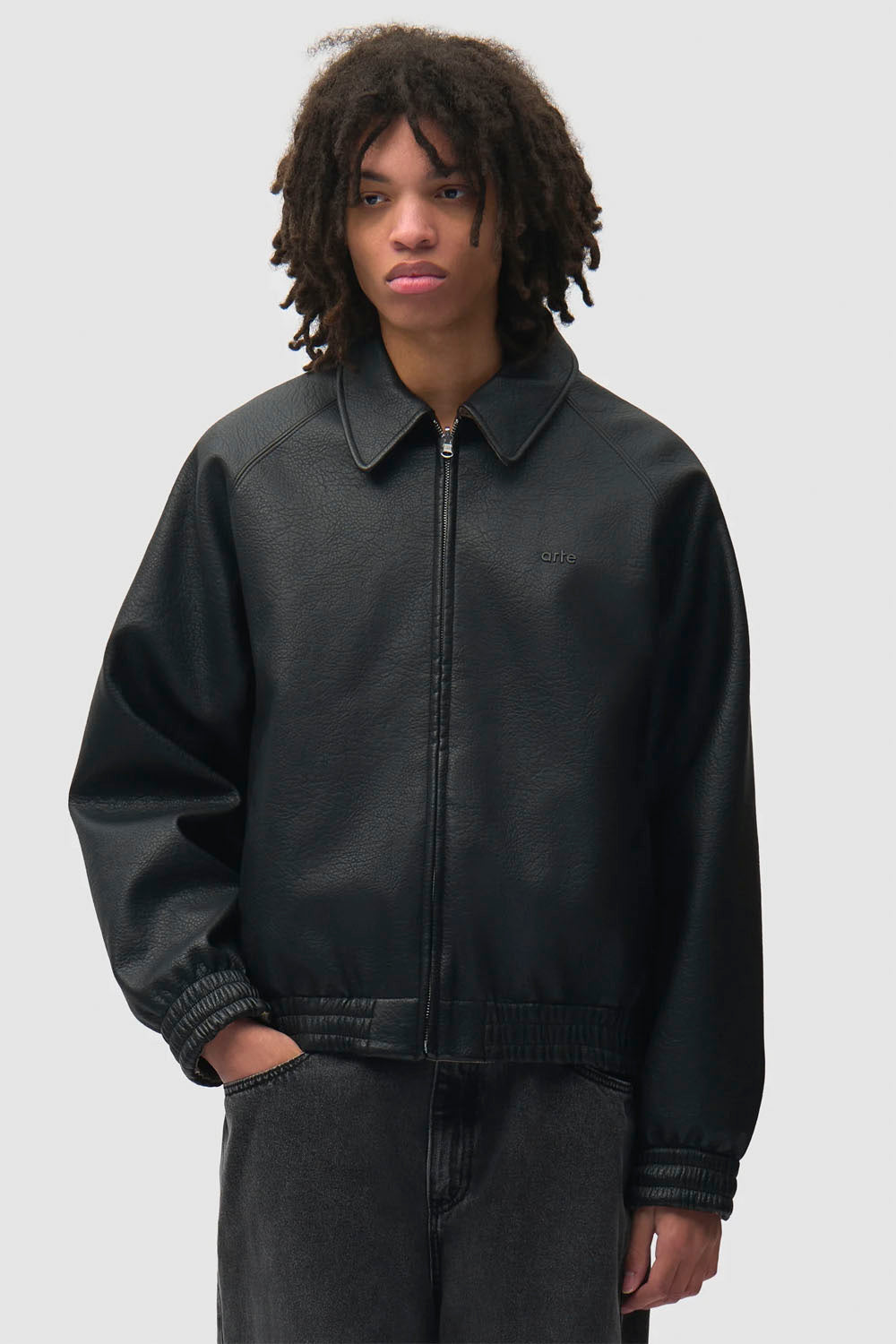 A model with curly dark hair wearing a black leather zip-up jacket and dark gray jeans, standing against a light gray studio background.