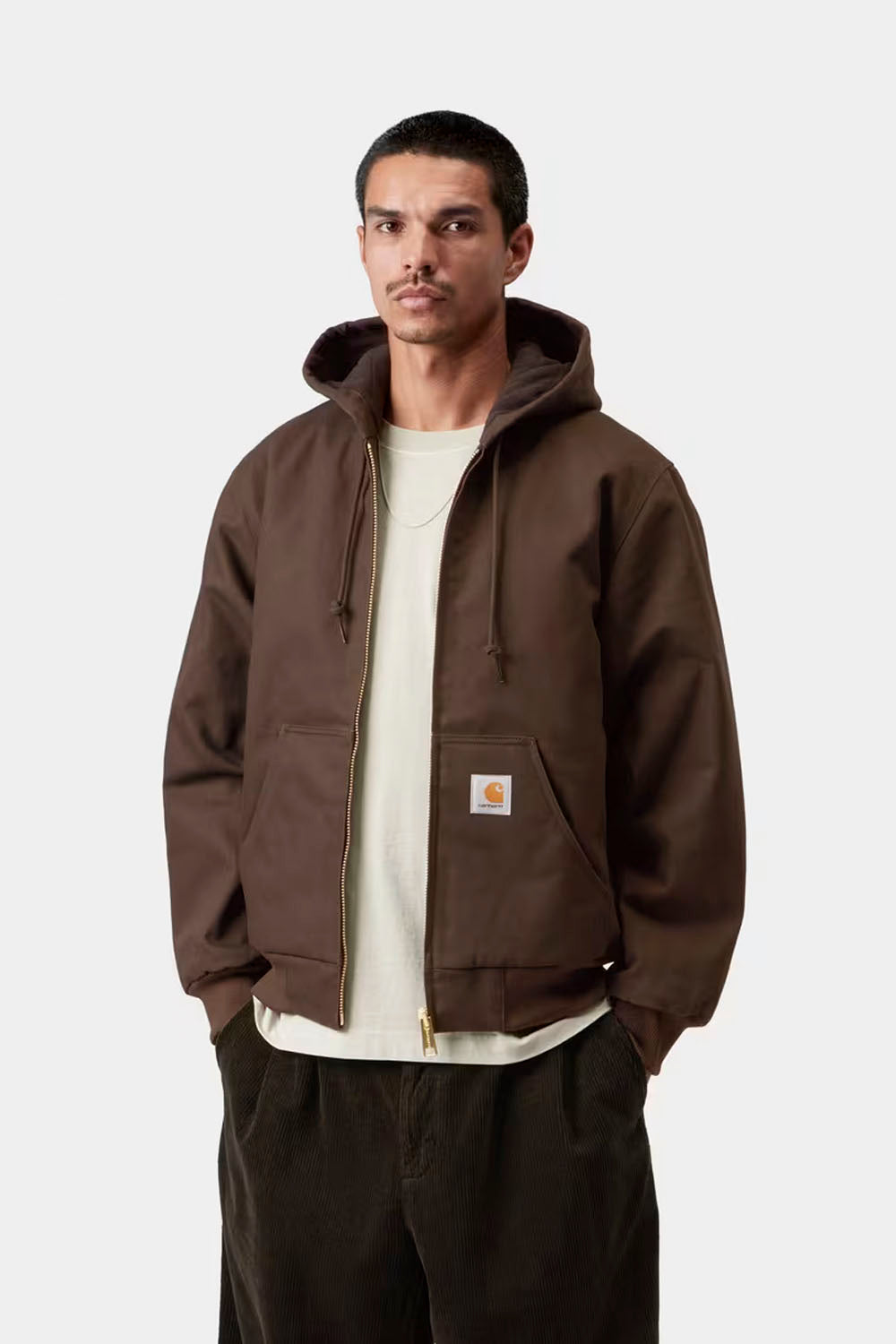 A man wearing a brown Carhartt hooded jacket over a cream t-shirt and dark pants, standing against a light gray studio background. The Carhartt logo patch is visible on the front pocket.