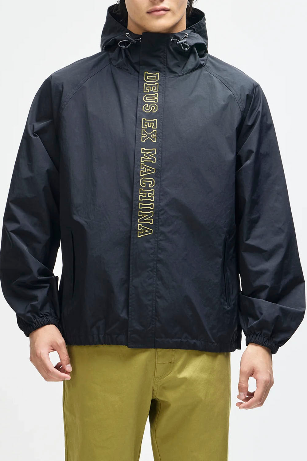 A man wearing a black hooded windbreaker jacket with 'DEUS EX MACHINA' printed vertically in gold on the front, paired with olive green pants, against a plain white background.