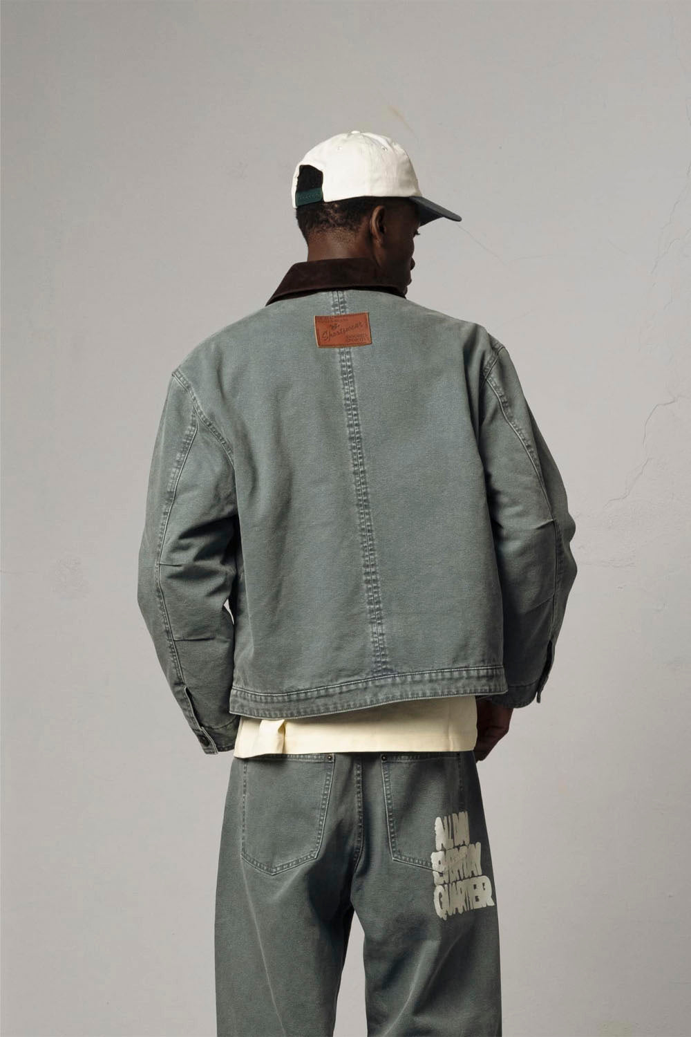 A model shown from the back wearing a muted gray-green canvas jacket with a brown collar, paired with matching gray-green pants and a white cap, against a plain gray studio background.