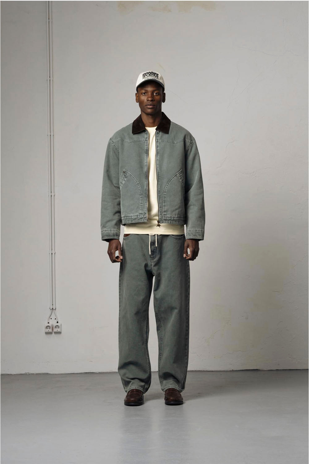 A model stands in a minimalist studio setting wearing a light gray denim jacket with a dark brown corduroy collar, paired with matching gray sweatpants, a cream t-shirt, and a white baseball cap.