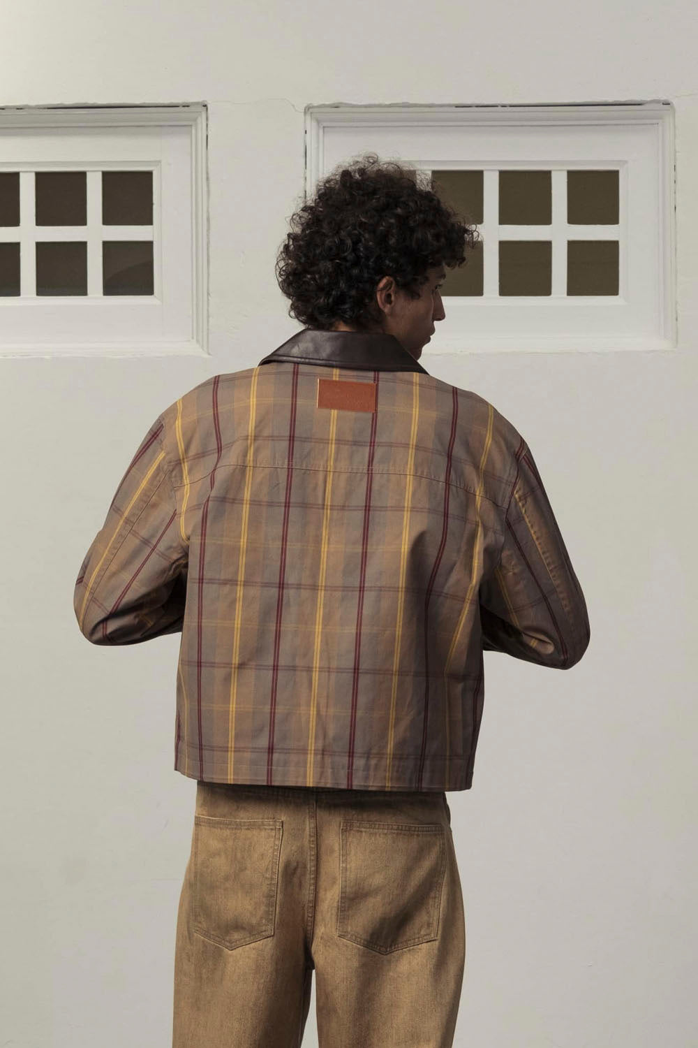 A man viewed from behind wearing a brown and yellow plaid cropped jacket with dark brown leather shoulder panels and a brown leather patch on the back, paired with brown trousers, standing against a white wall with two window frames.