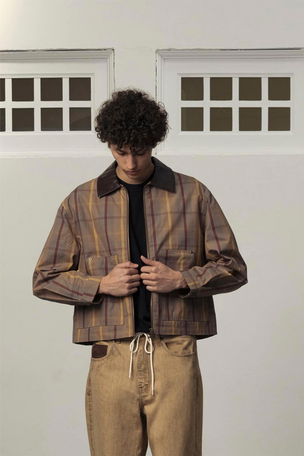 A man wearing a brown and beige plaid button-up jacket with a dark collar, styled over a black shirt and tan drawstring jeans, standing against a white wall with two window frames.