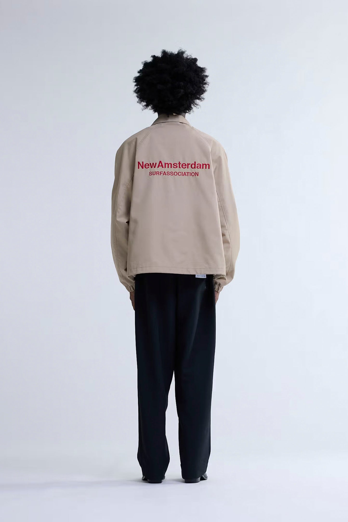 Person wearing New Amsterdam Surf Association Coach Jacket in Sand with 'New Amsterdam' text on the back against a white background