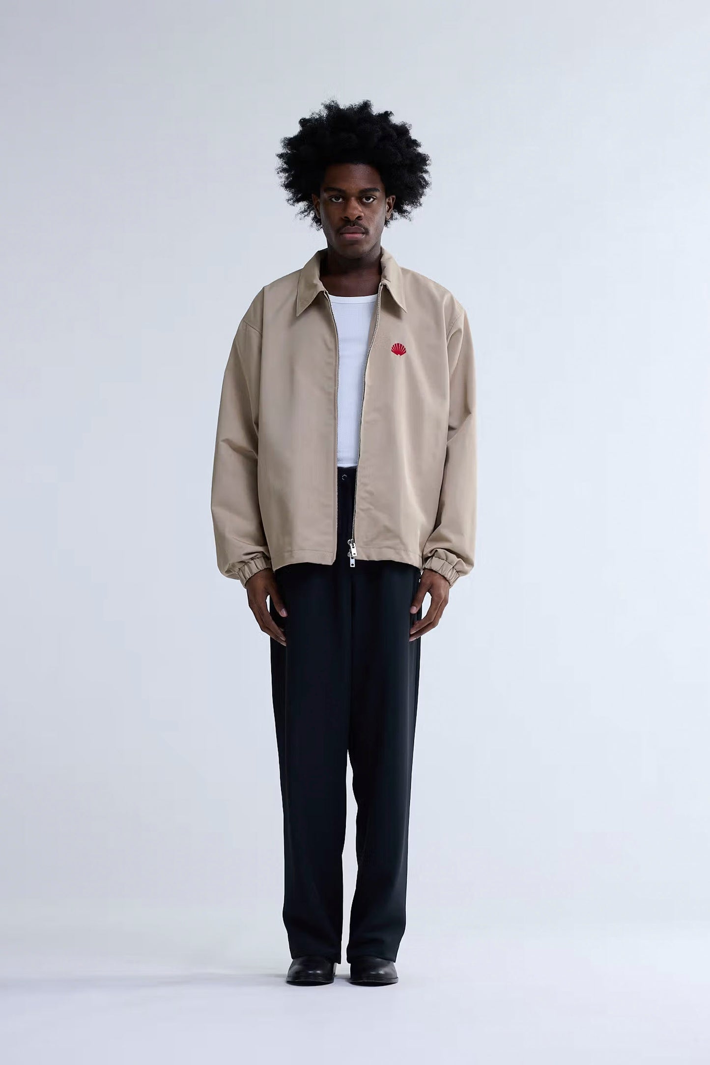 Man wearing New Amsterdam Surf Association Coach Jacket in Sand over a white shirt with a red pin, standing against a plain background