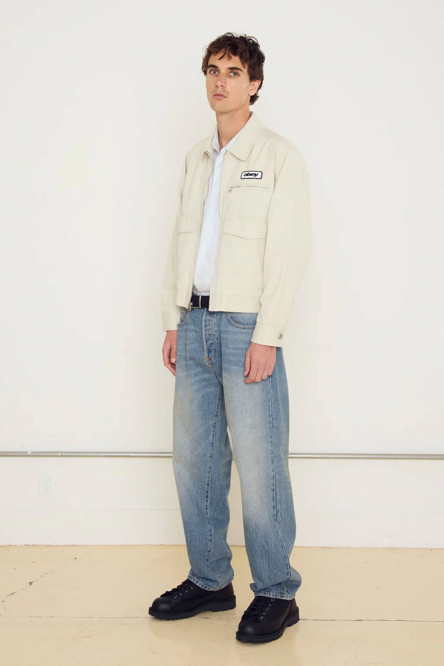 Man wearing a Beige OBEY Walter Work Jacket , white shirt, blue jeans, and black shoes on a white background