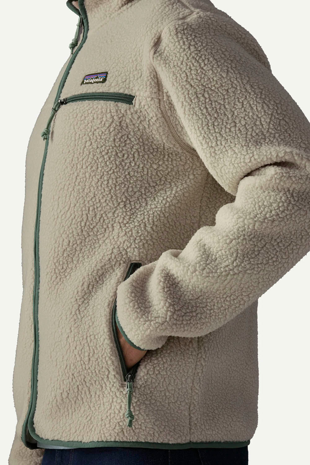 Close-up of a Patagonia fleece jacket in a light beige/cream color with dark green zipper and trim details, showing the chest pocket and side profile.