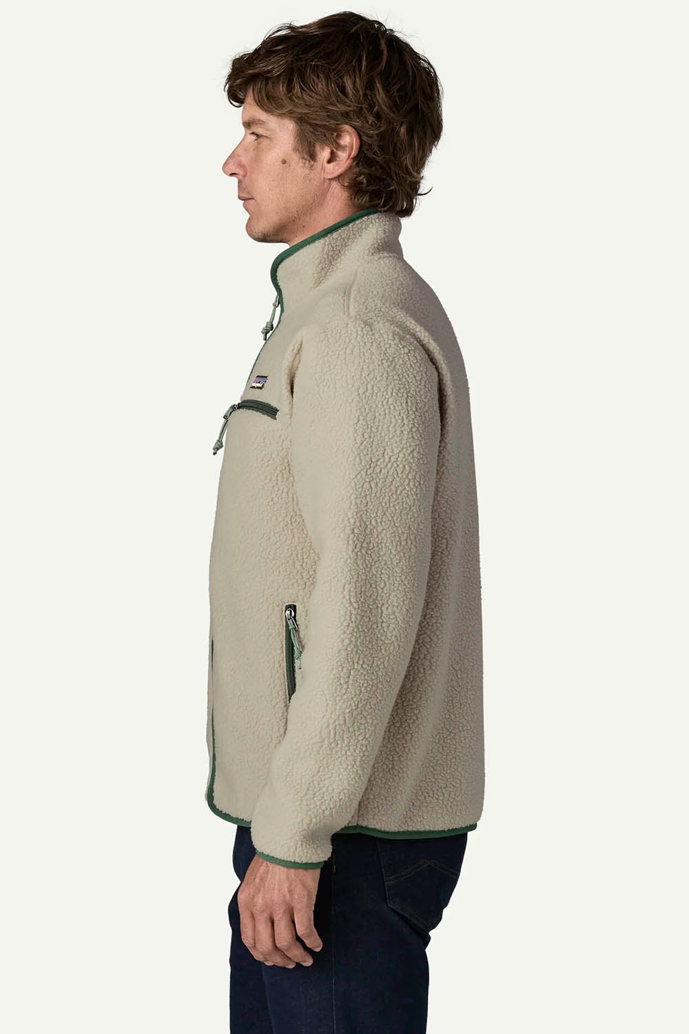 A man in profile wearing a light beige sherpa fleece pullover jacket with dark green trim and zipper details, paired with dark blue jeans, against a plain off-white background.