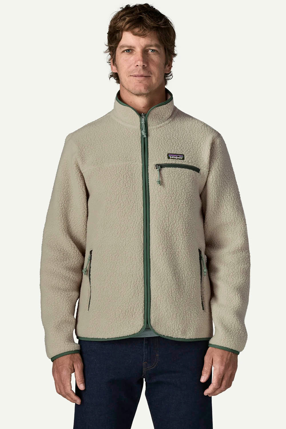 A man wearing a beige Patagonia fleece jacket with dark green trim and zipper details, paired with dark blue jeans, against a light gray studio background.
